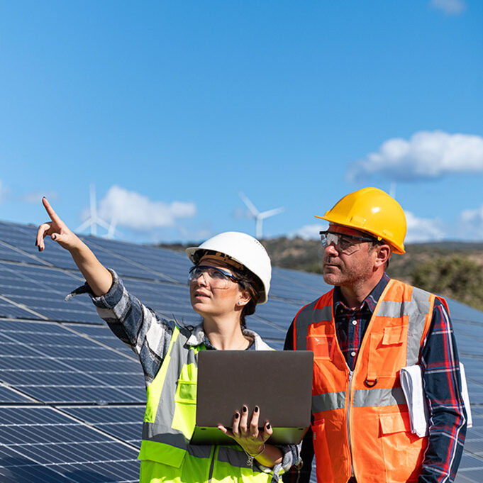 A man and a woman wearing safety helmets and high-visibility waistcoats stand in front of solar panels. The woman has a laptop in her hand and is pointing into the distance with her other hand.