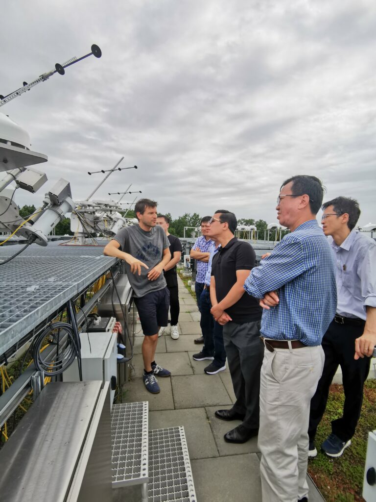 The delegation stands on the roof platform of the German Weather Service (DWD) in Lindenberg, Thuringia. The DWD employee explains how pyranometers work, which can be seen in the background. These pyranometers measure the diffuse irradiation of the sun and track its course, as they are mounted on trackers.