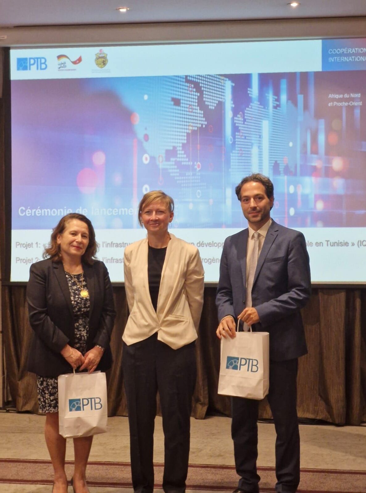 Two women and a man are standing in front of a screen with a blue world map. The woman on the left and the man on the right are each holding a white bag in their hands.