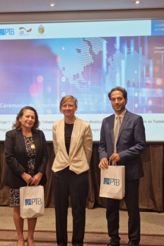 Two women and a man are standing in front of a screen with a blue world map. The woman on the left and the man on the right are each holding a white bag in their hands.