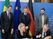 A woman and a man sitting at a desk, signing documents and laughing. Behind them, three men are standing in front of three flags.