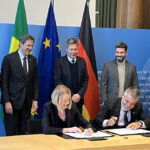 A woman and a man sitting at a desk, signing documents and laughing. Behind them, three men are standing in front of three flags.