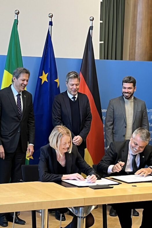 A woman and a man sitting at a desk, signing documents and laughing. Behind them, three men are standing in front of three flags.