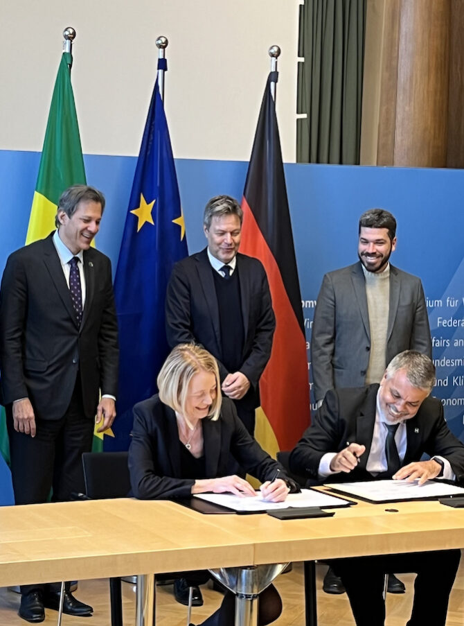 A woman and a man sitting at a desk, signing documents and laughing. Behind them, three men are standing in front of three flags.