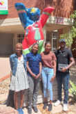 Four Namibian Master’s students standing outside the University of Namibia. The Berlin Bear statue behind them has raised its paws to wish them good luck.