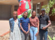 Four Namibian Master’s students standing outside the University of Namibia. The Berlin Bear statue behind them has raised its paws to wish them good luck.