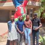 Four Namibian Master’s students standing outside the University of Namibia. The Berlin Bear statue behind them has raised its paws to wish them good luck.