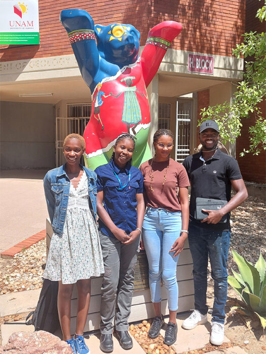 Four Namibian Master’s students standing outside the University of Namibia. The Berlin Bear statue behind them has raised its paws to wish them good luck.
