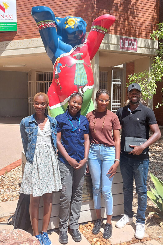 Four Namibian Master’s students standing outside the University of Namibia. The Berlin Bear statue behind them has raised its paws to wish them good luck.