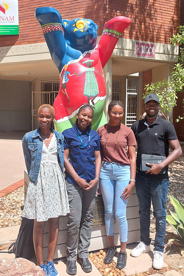 Four Namibian Master’s students standing outside the University of Namibia. The Berlin Bear statue behind them has raised its paws to wish them good luck.