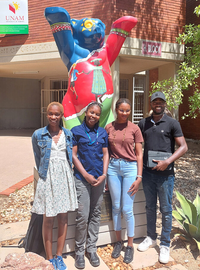 Four Namibian Master’s students standing outside the University of Namibia. The Berlin Bear statue behind them has raised its paws to wish them good luck.