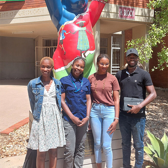 Four Namibian Master’s students standing outside the University of Namibia. The Berlin Bear statue behind them has raised its paws to wish them good luck.