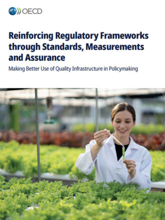 A woman wearing a lab coat is standing inside of a greenhouse. She is in front of young plants on long tables and is holding a pipette above a test tube.