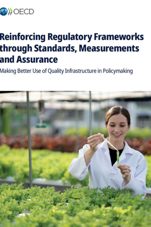 A woman wearing a lab coat is standing inside of a greenhouse. She is in front of young plants on long tables and is holding a pipette above a test tube.