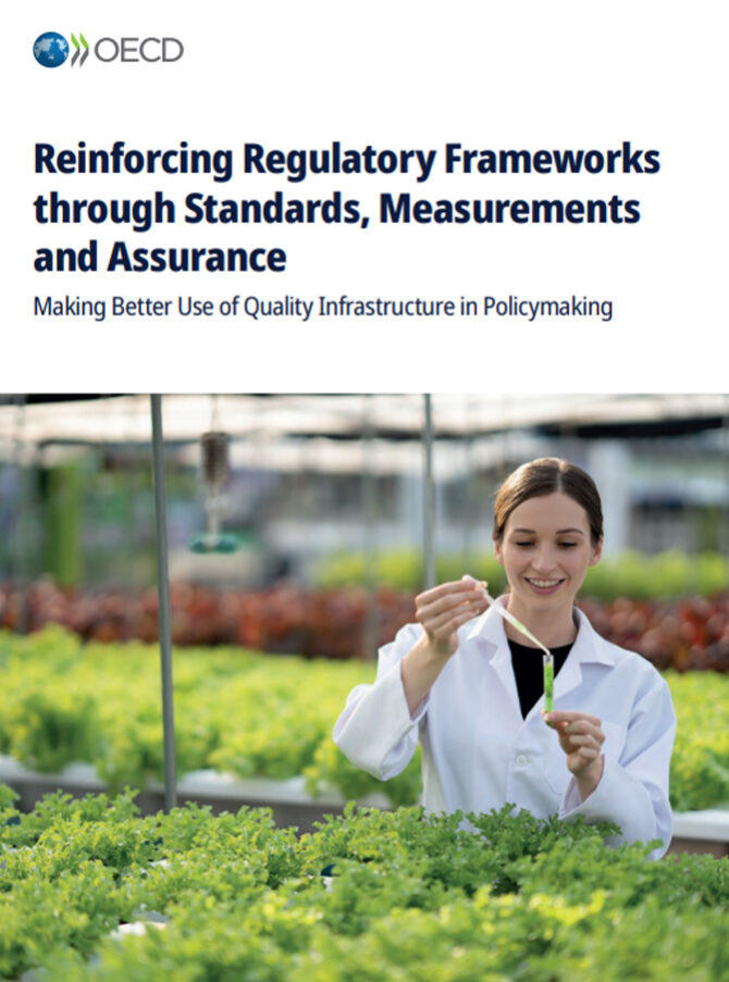 A woman wearing a lab coat is standing inside of a greenhouse. She is in front of young plants on long tables and is holding a pipette above a test tube.