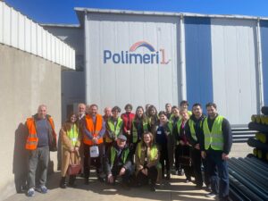 A group of men and women wearing neon safety vests are posing in front of the Polimeri 1 building.