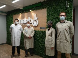 Eine Frau und drei Männer tragen Laborkittel und Gesichtsmasken und stehen vor einer Wand, auf der das Wort „Analab“ steht.