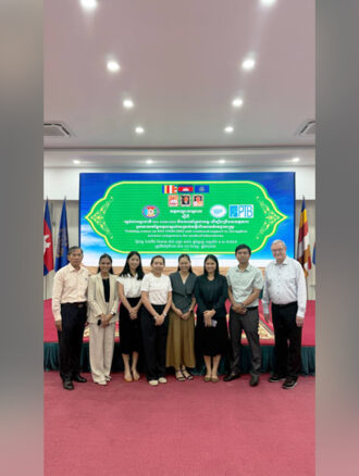Alternativtext: Five women and three men standing in front of a stage and a large banner in a conference room in Cambodia ©GDAC