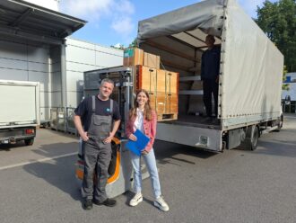 A man and a woman are standing in front of a truck loaded with wooden boxes full of equipment to be sent to Ukraine.
