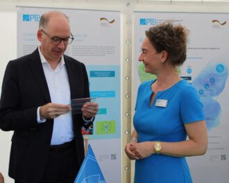 BMZ-Staatssekretär Niels Annen und Dr. Barbara Siegmund stehen vor zwei Roll-ups. Er hält eine Wellplatte aus Kunststoff in den Händen. © PTB/Ella Jones