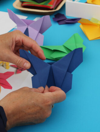 Zwei Hände, die einen dunkelblauen Schmetterling aus gefaltetem Papier halten. Im Hintergrund sind weitere gefaltete Schmetterlinge und Papierstücke auf einer blauen Tischdecke zu sehen.