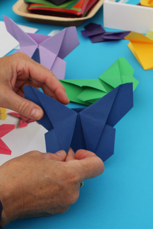 Zwei Hände, die einen dunkelblauen Schmetterling aus gefaltetem Papier halten. Im Hintergrund sind weitere gefaltete Schmetterlinge und Papierstücke auf einer blauen Tischdecke zu sehen.